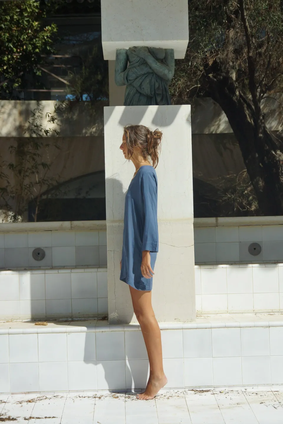Femme en robe de coton bleu de profil contre un pilier blanc au bord d'un bassin, chignon défait et pieds nus, à Porquerolles.