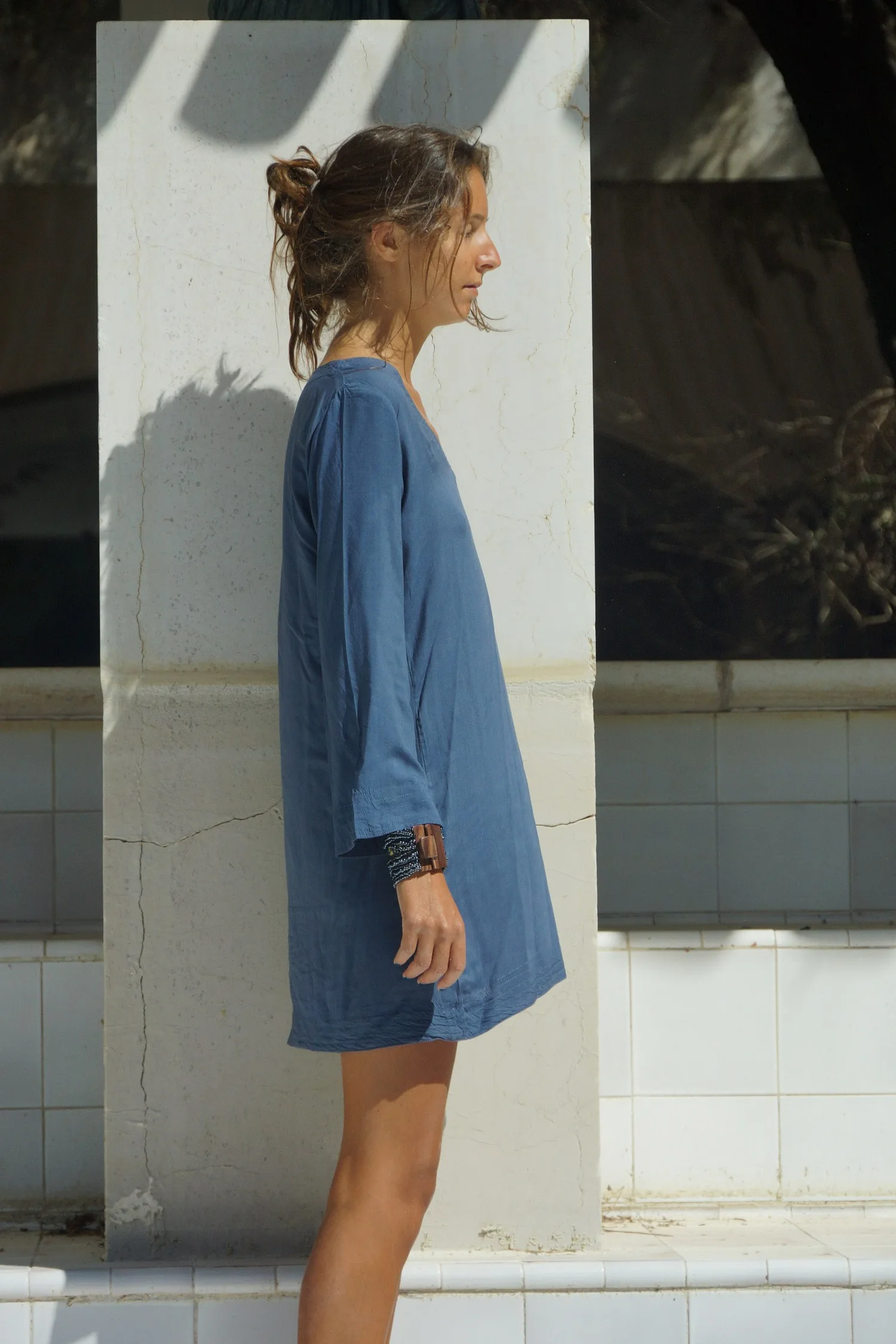 Femme de profil en robe de coton bleu à manches longues, bracelet de bois, adossée à un pilier blanc à Porquerolles.