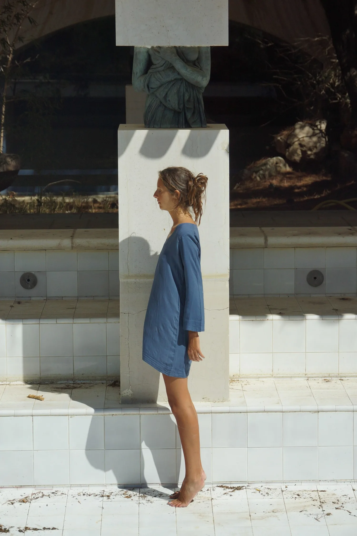 Femme en robe de coton bleu de profil contre un pilier surmonté d'une sculpture, bassin carrelé blanc, à Porquerolles.