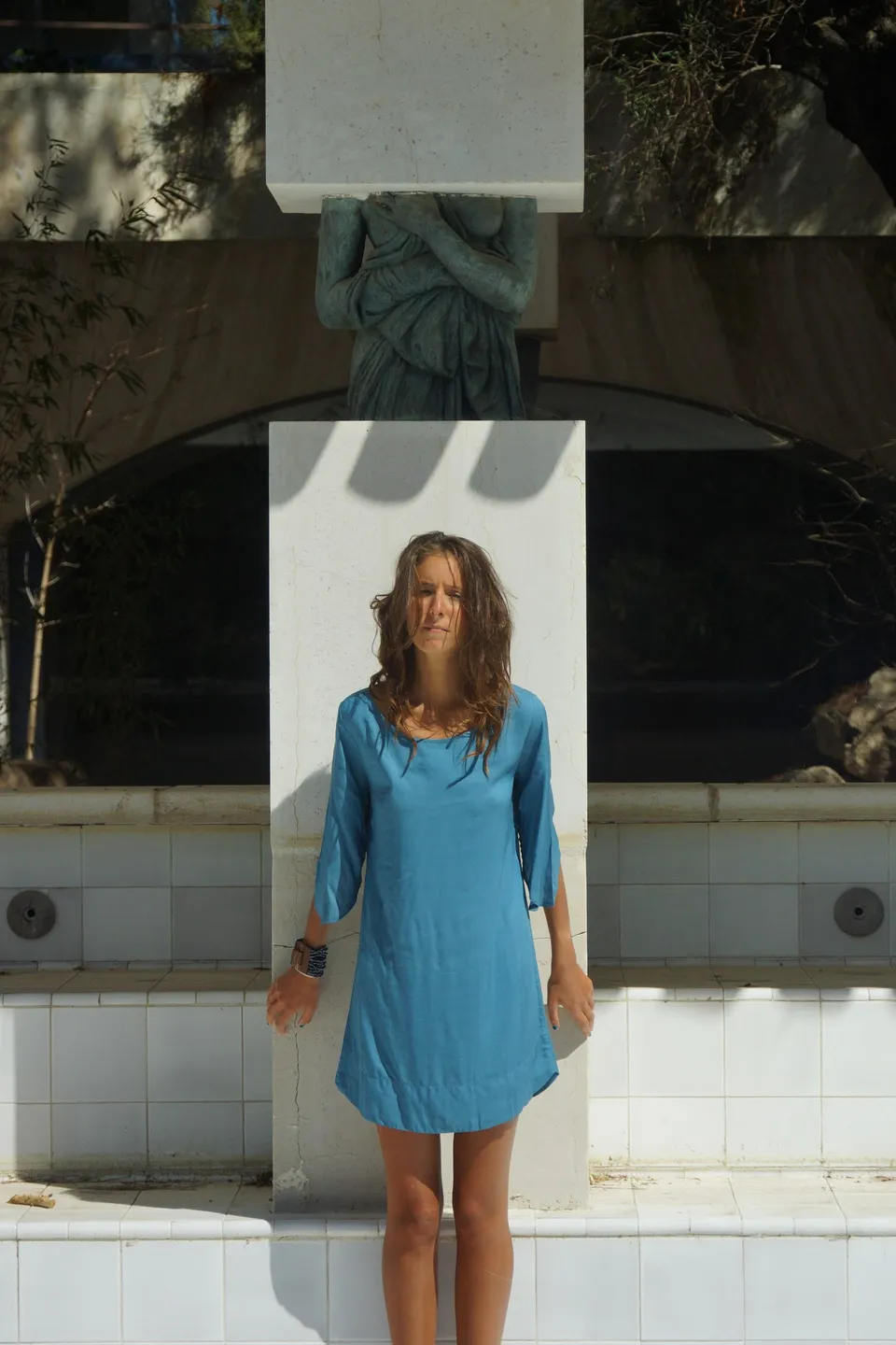 Femme en tunique de lin turquoise debout de face devant un pilier surmonté d'une sculpture, à Porquerolles.