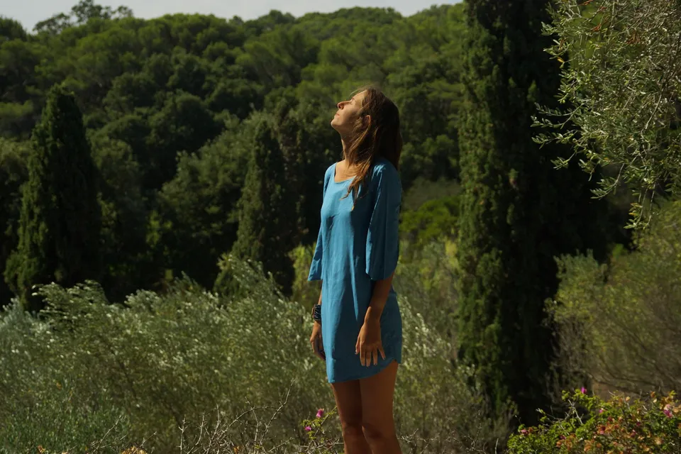 Femme en tunique de lin turquoise debout dans le maquis, visage renversé vers le ciel, cyprès et oliviers à Porquerolles.