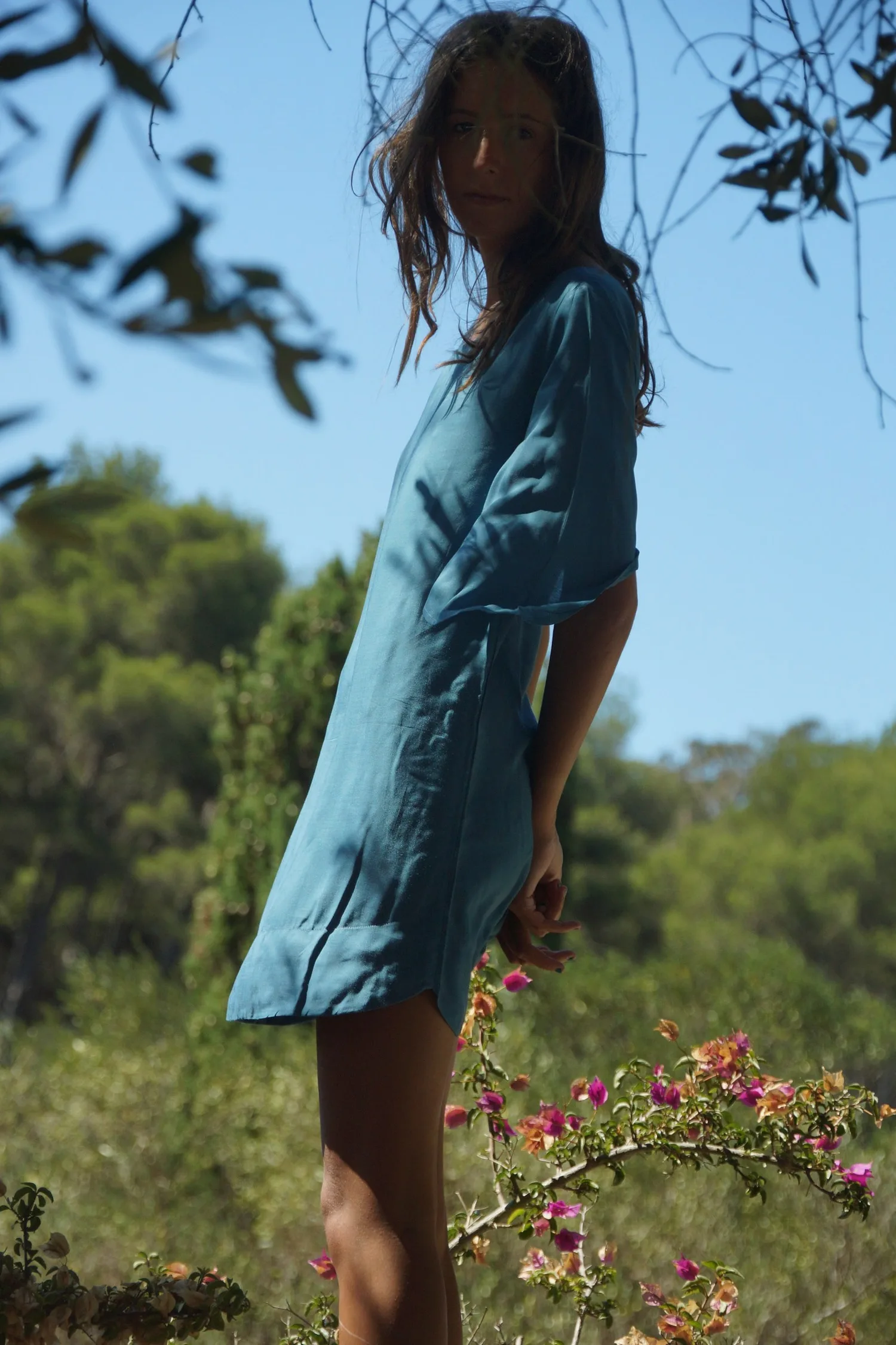 Femme en tunique de lin turquoise entre les branches d'un olivier, bougainvillier rose au premier plan, à Porquerolles.