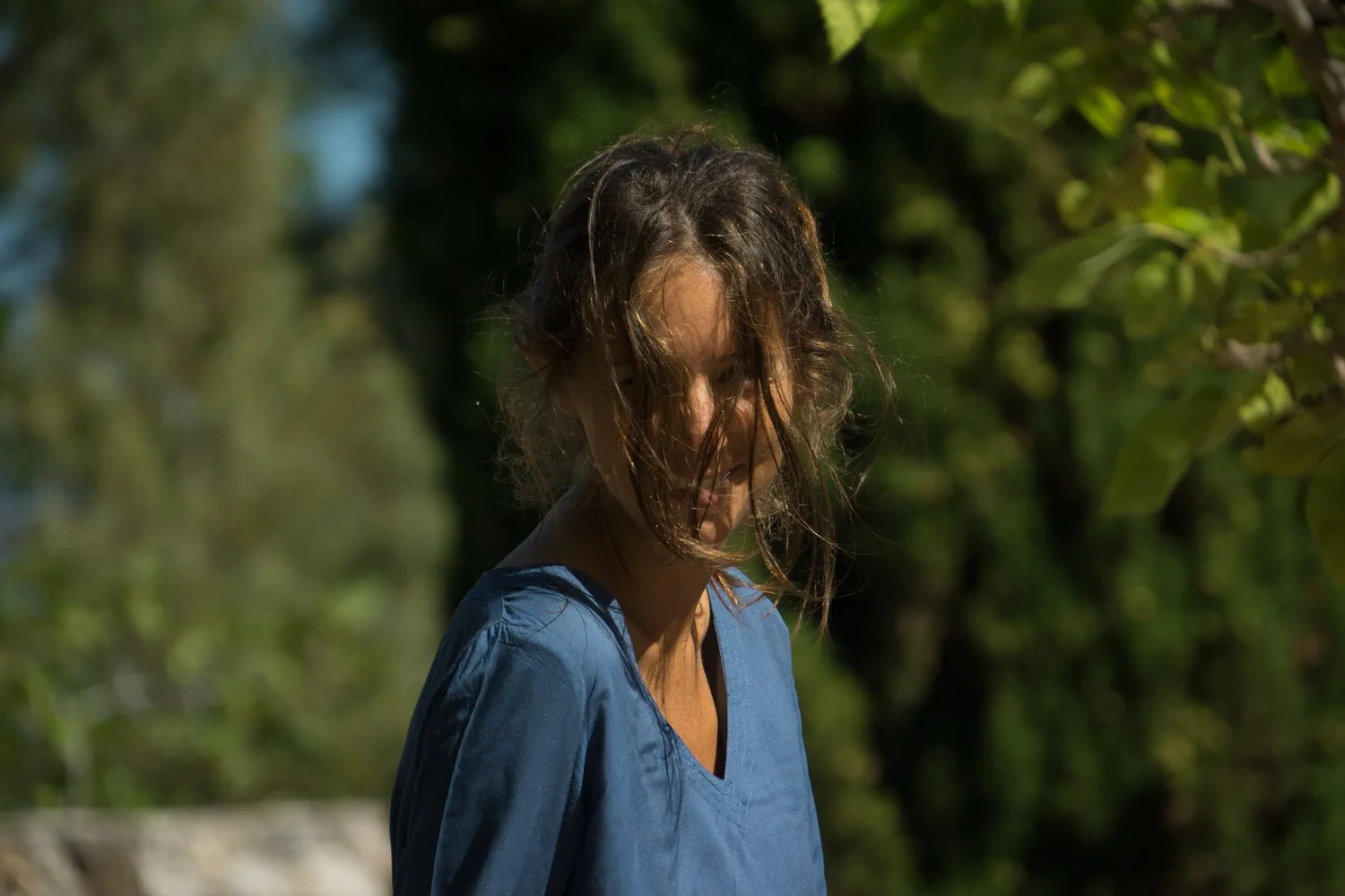 Portrait de profil d'une femme en robe de coton bleu, cheveux dans le visage, cyprès en arrière-plan à Porquerolles.