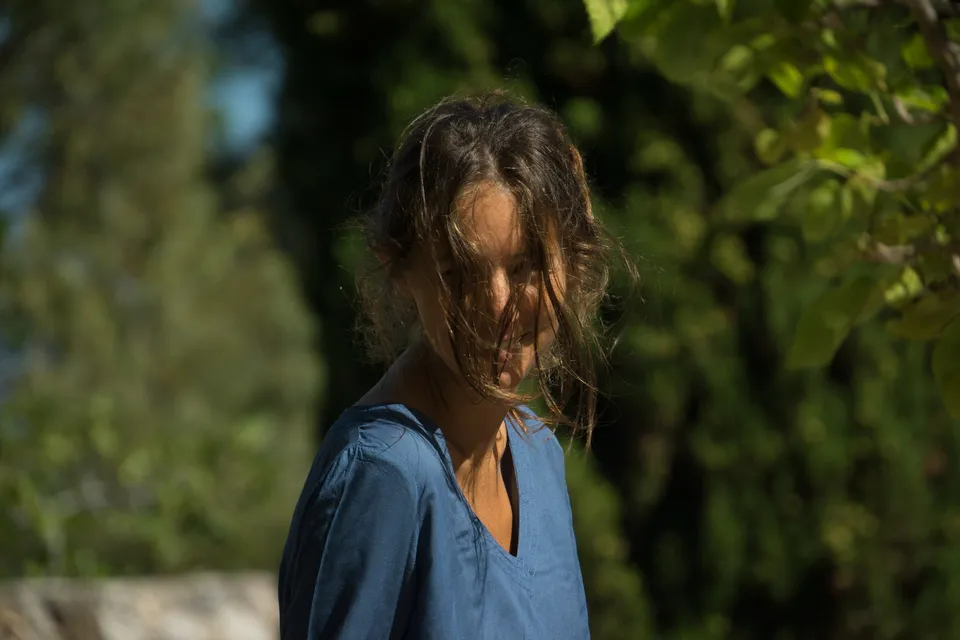Portrait de profil d'une femme en robe de coton bleu, cheveux dans le visage, cyprès en arrière-plan à Porquerolles.