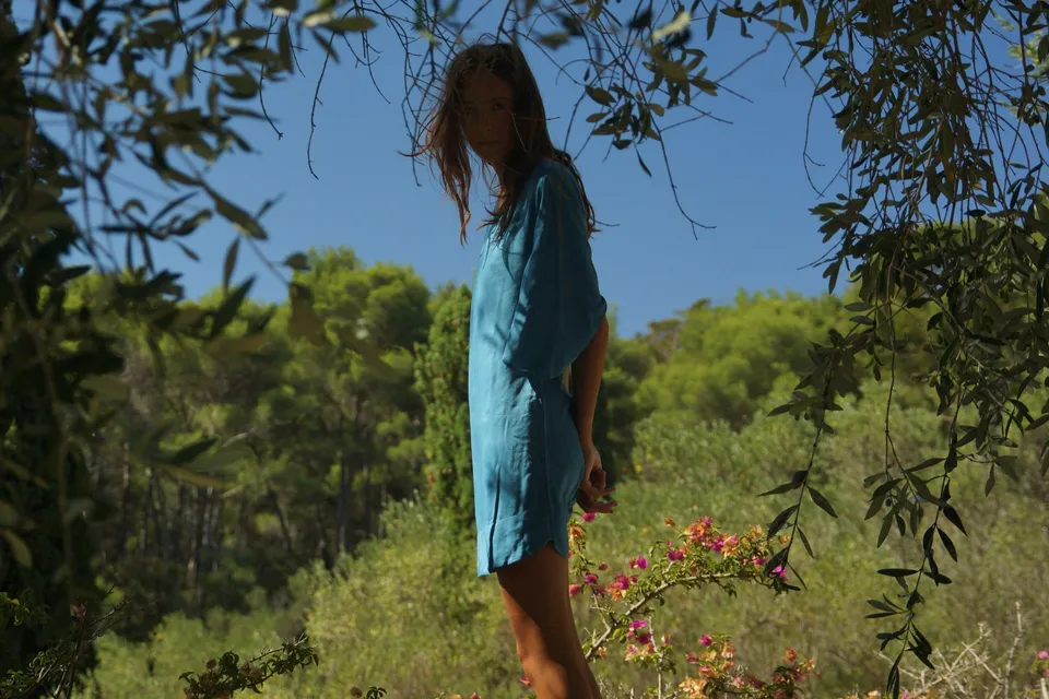Femme en tunique de lin turquoise debout dans le maquis, regard par-dessus l'épaule, branches d'olivier en cadre, à Porquerolles.