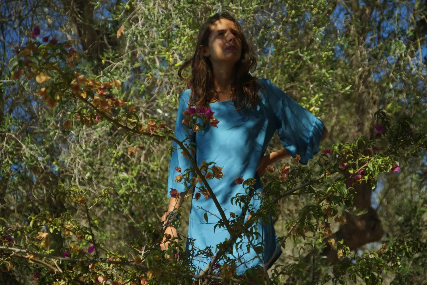 Femme en tunique de lin turquoise debout parmi des branches fleuries, main sur la hanche et regard levé, à Porquerolles.