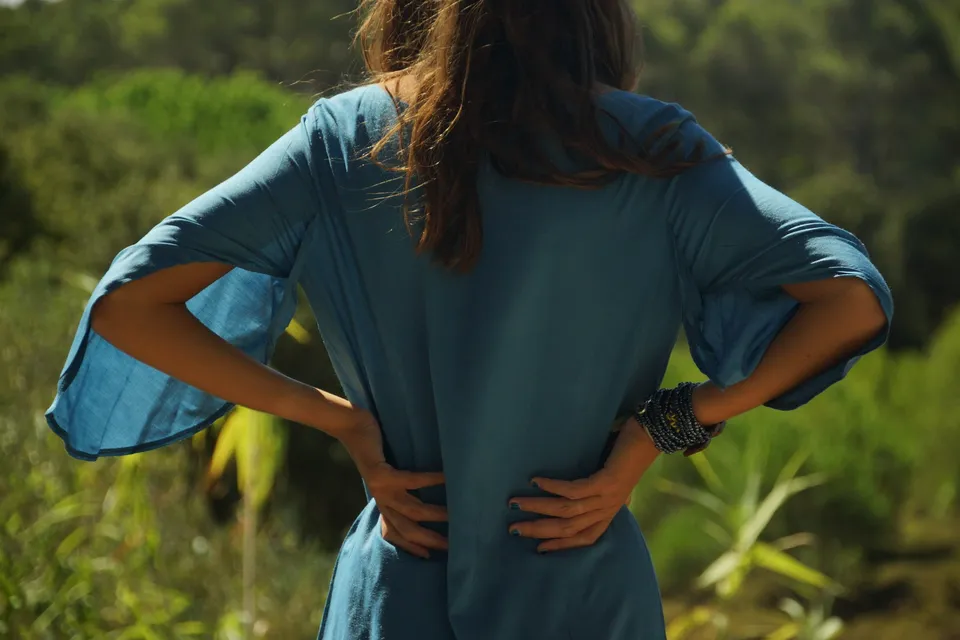 Dos d'une femme en tunique de lin turquoise, mains aux hanches et manches évasées, maquis flou derrière, à Porquerolles.