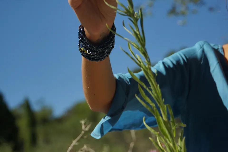 Détail d'une main touchant une tige verte, manchette de perles noires et manche turquoise, sur fond de ciel bleu à Porquerolles.