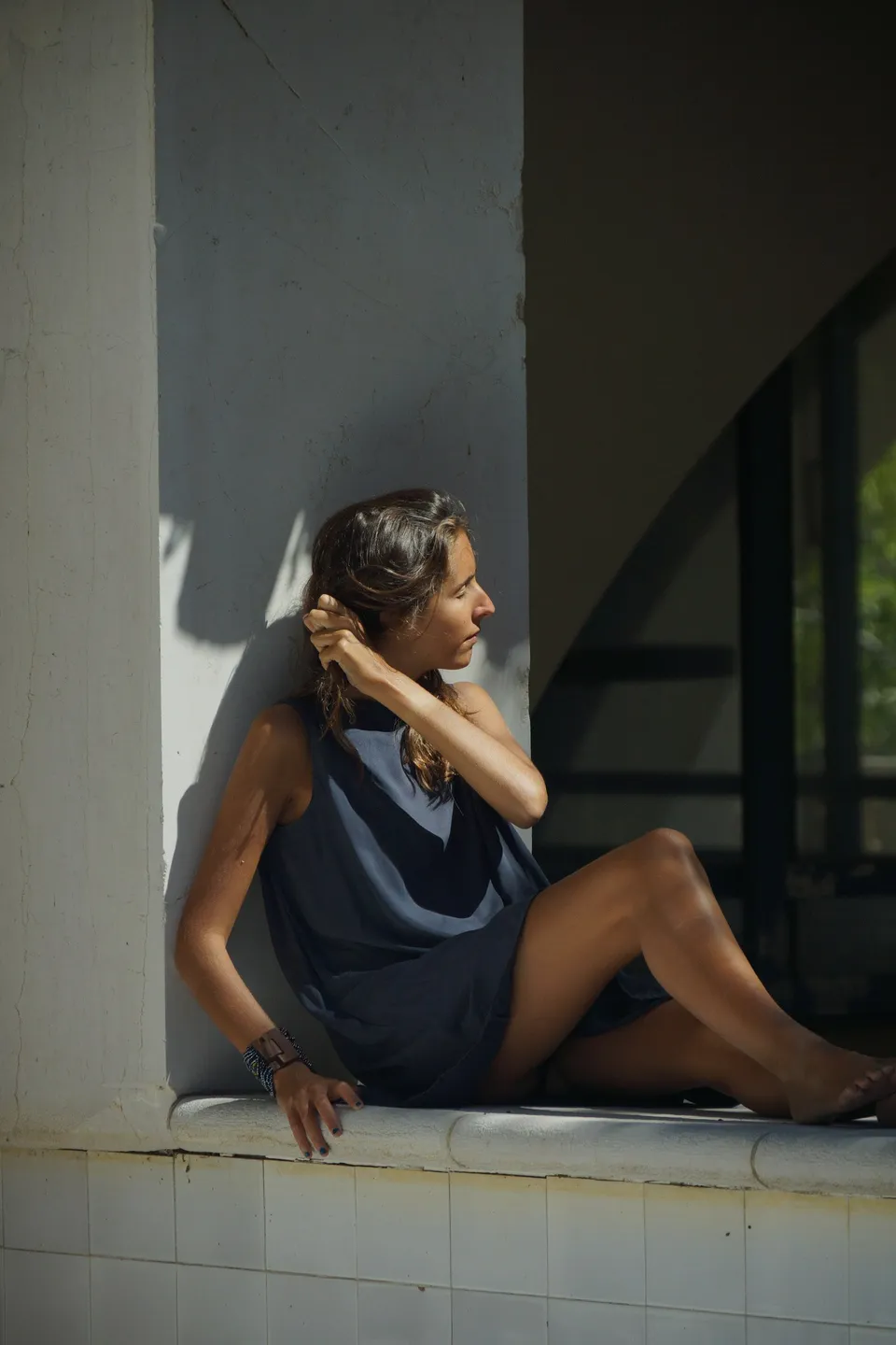 Femme en robe drapée gris-bleu sans manches assise contre un pilier, main dans les cheveux, à Porquerolles.