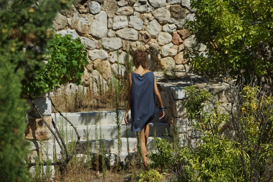 Femme de dos en robe drapée gris-bleu devant un mur de pierre sèche, manchette au poignet, à Porquerolles.