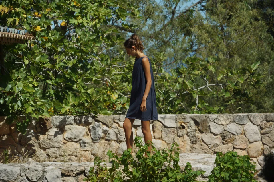 Femme en robe drapée gris-bleu debout sur un muret sous un figuier, regard baissé, à Porquerolles.