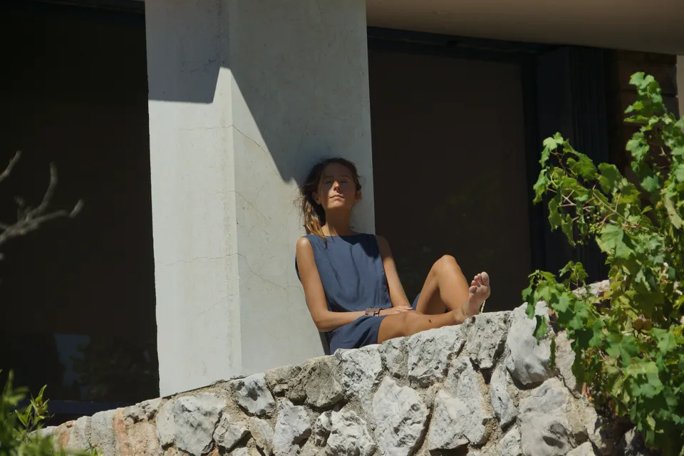 Femme en robe gris-bleu assise sur un muret contre un pilier, jambes repliées et visage au soleil, vigne à Porquerolles.