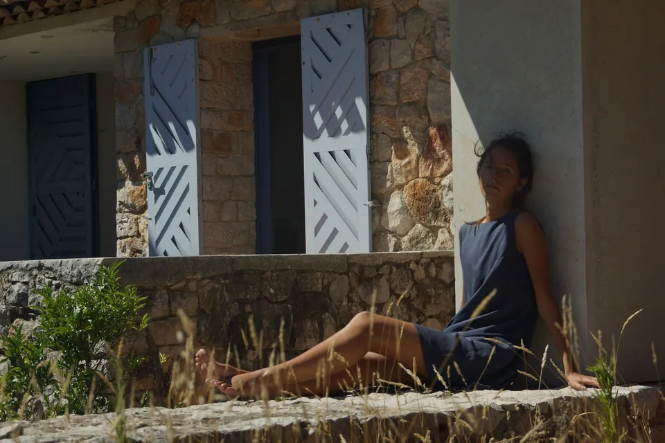 Femme en robe gris-bleu assise contre le mur d'une maison de pierre à volets clairs, jambes tendues dans les herbes sèches, à Porquerolles.