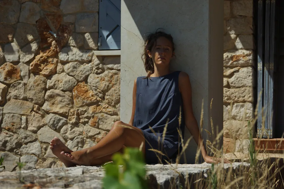 Femme en robe gris-bleu sans manches assise au pied d'un mur de pierre à volets clairs, jambes tendues, à Porquerolles.