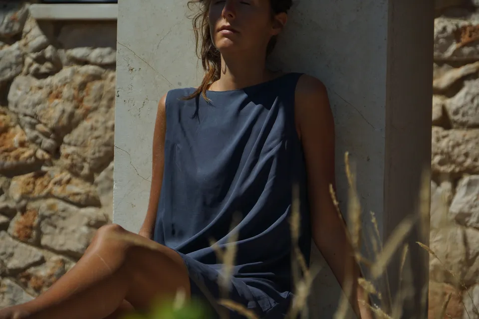 Femme en robe drapée gris-bleu adossée à un pilier, yeux fermés au soleil, mur de pierre derrière, à Porquerolles.
