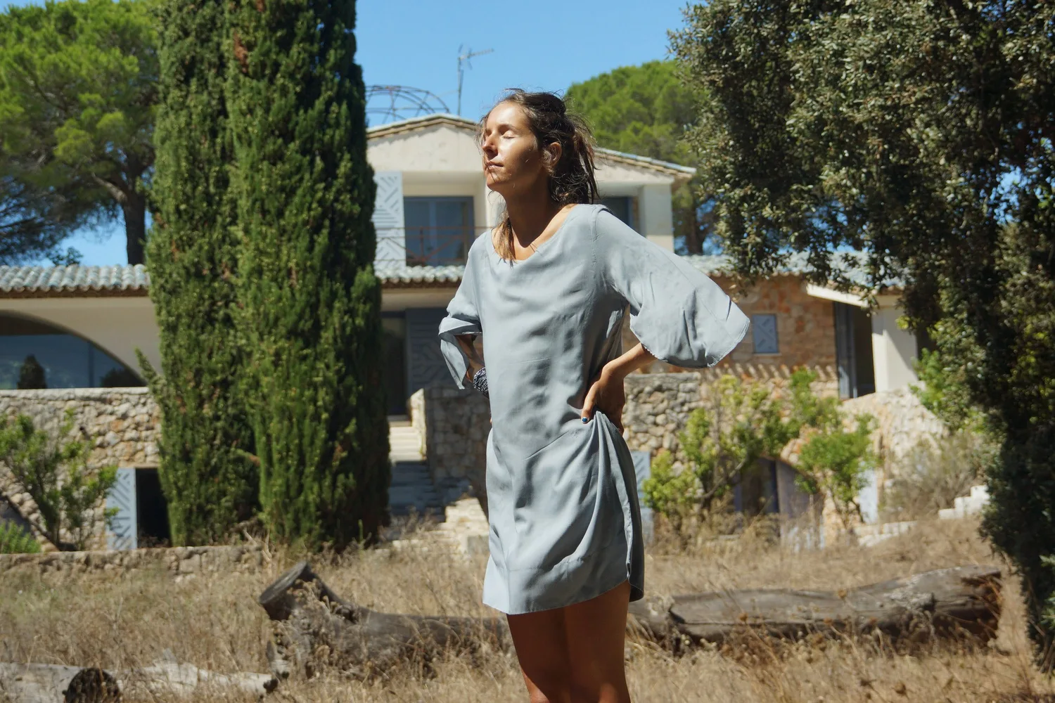 Femme en robe de lin gris clair à manches évasées debout devant une maison de pierre et des cyprès à Porquerolles.