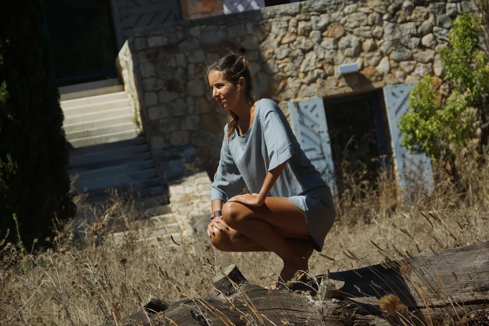 Femme en robe de lin gris clair accroupie dans les herbes sèches devant une maison de pierre à Porquerolles.
