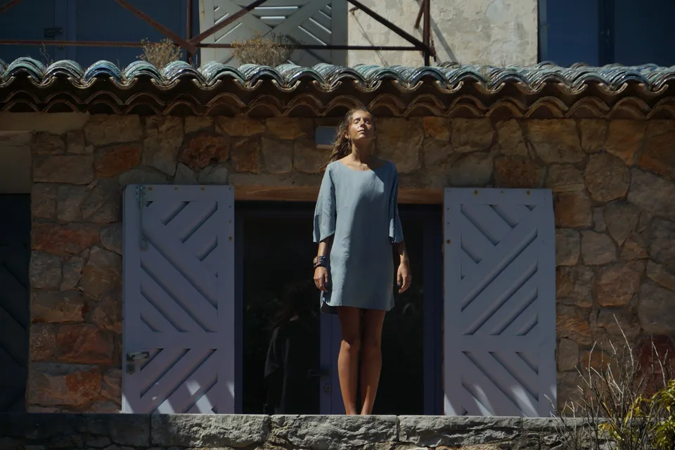 Femme en robe de lin gris clair debout sur un seuil entre deux volets lilas, façade de pierre, à Porquerolles.