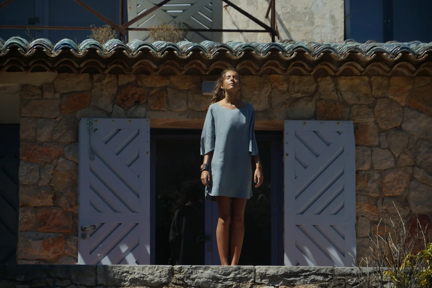 Femme en robe de lin gris clair debout sur un seuil entre deux volets lilas, façade de pierre, à Porquerolles.
