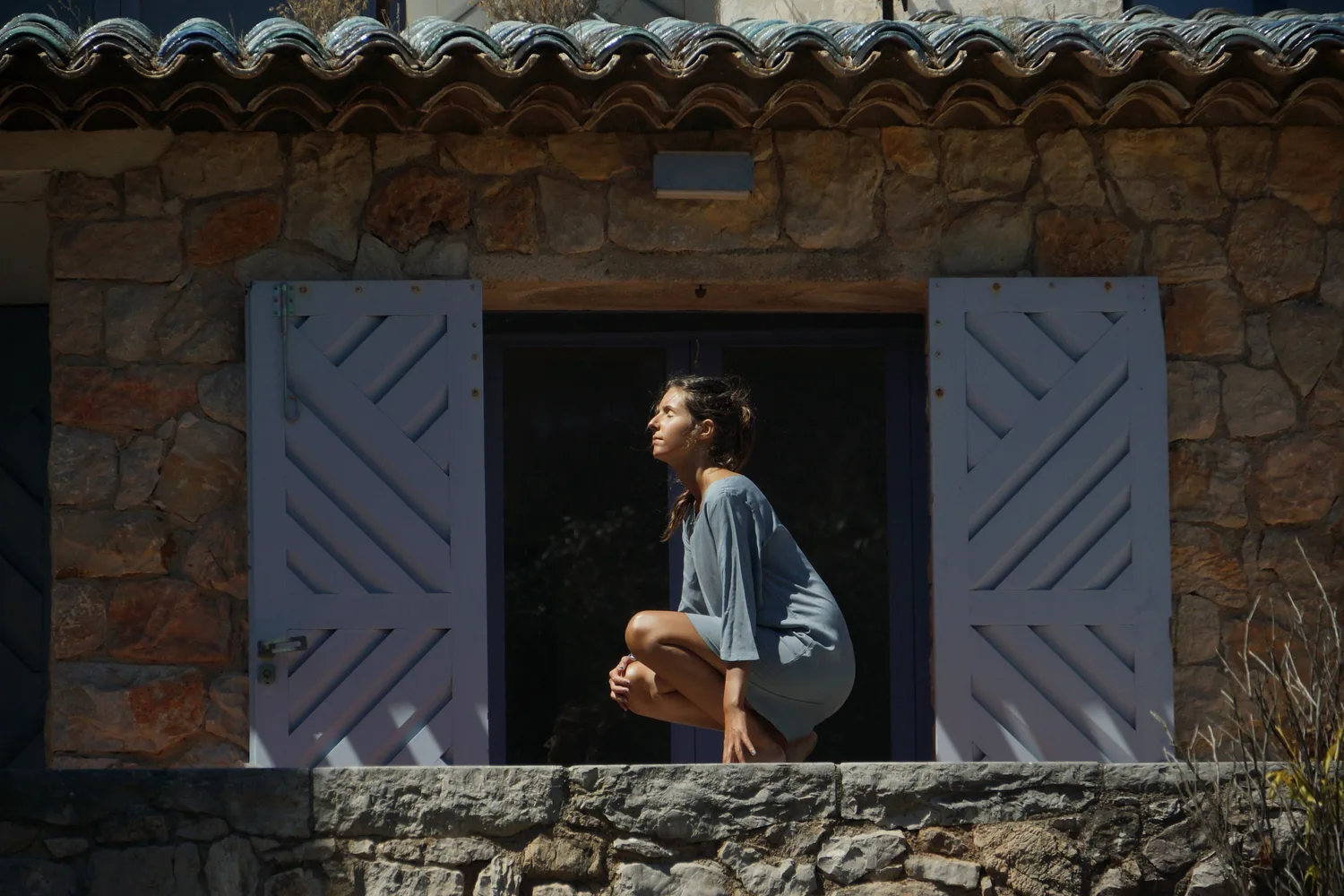 Femme accroupie sur le seuil d'une maison de pierre entre deux volets lilas, profil tourné vers le soleil, à Porquerolles.