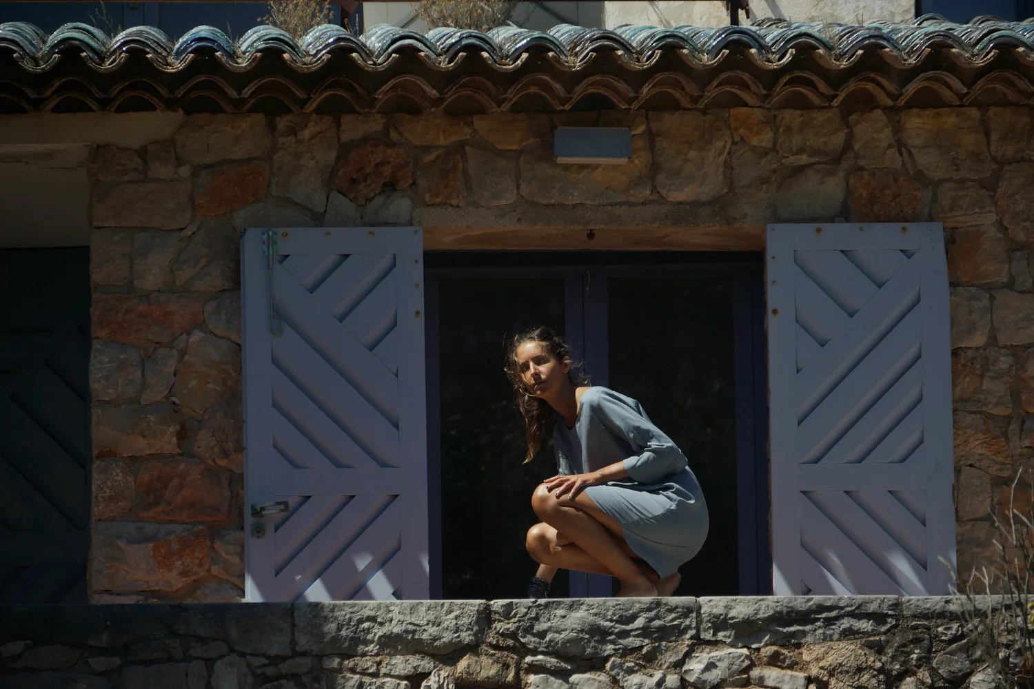 Femme accroupie dans l'embrasure sombre d'une maison de pierre aux volets lilas, regard vers l'objectif, à Porquerolles.