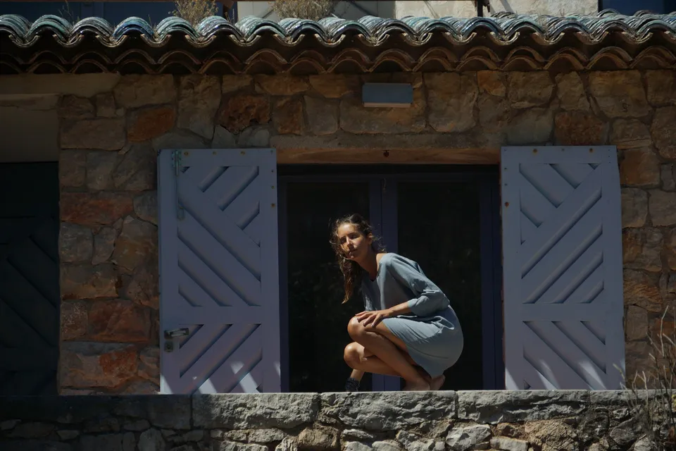 Femme accroupie dans l'embrasure sombre d'une maison de pierre aux volets lilas, regard vers l'objectif, à Porquerolles.