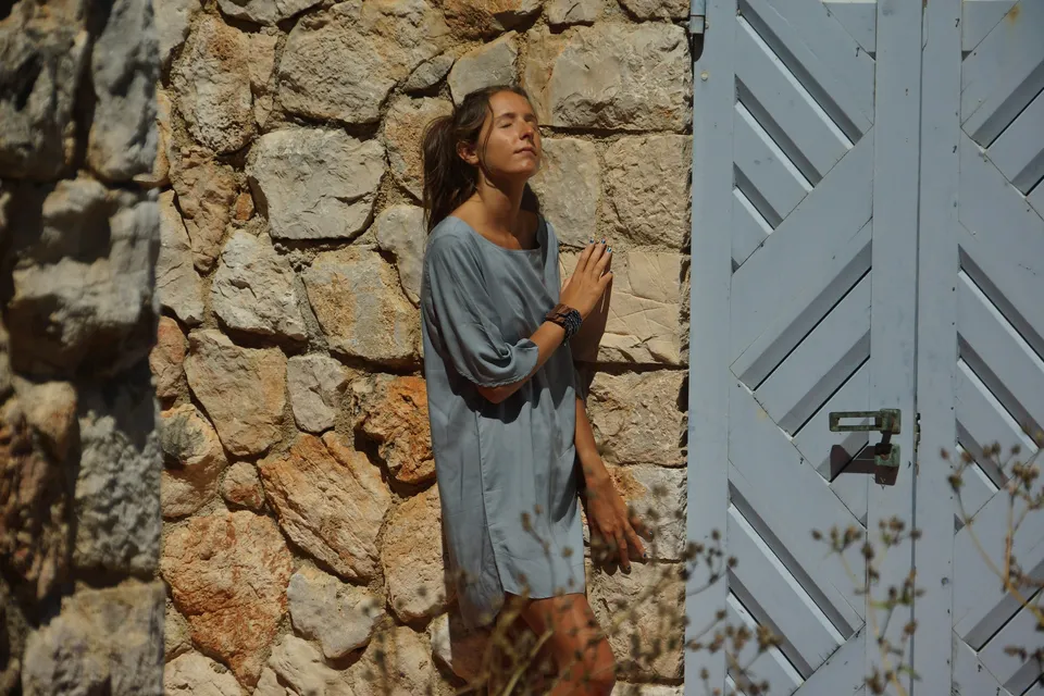 Femme en robe de voile gris-bleu adossée à un mur de pierre, yeux clos et mains repliées, près d'un volet blanc, à Porquerolles.