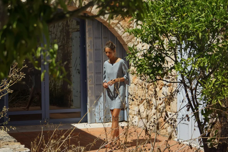 Femme en robe de voile gris-bleu marchant devant une porte voûtée bleue, branchages au premier plan, à Porquerolles.