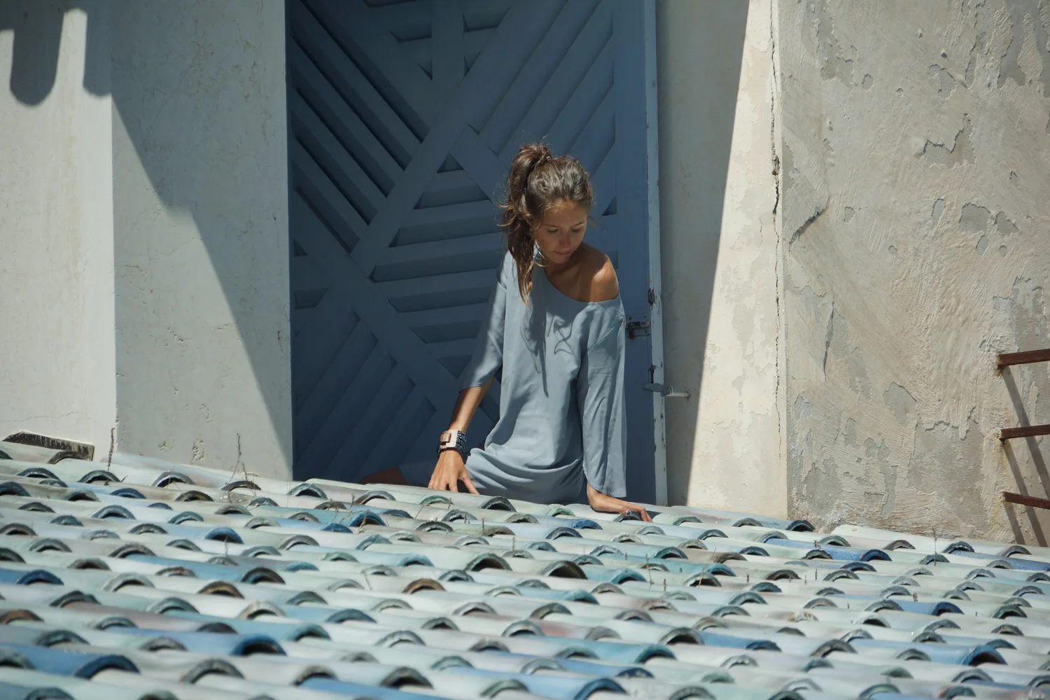 Femme en robe de lin gris-bleu, épaule nue, sur une terrasse au-dessus d'un toit de tuiles bleutées, à Porquerolles.