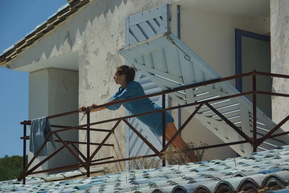 Femme en robe de lin bleu penchée à une rambarde de fer près d'un linge qui sèche, persiennes ouvertes, à Porquerolles.
