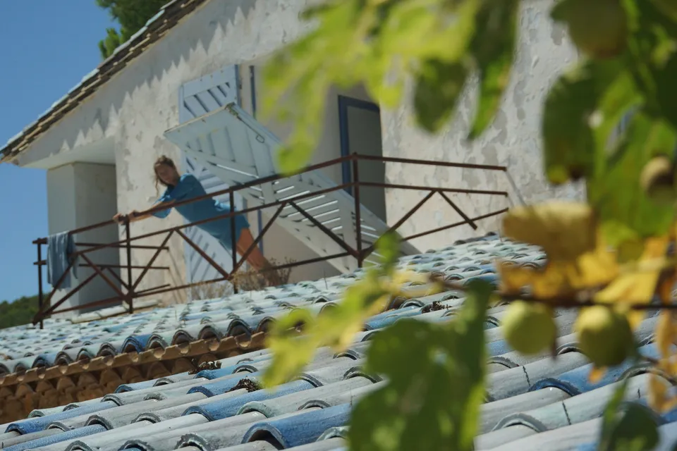 Femme en robe de coton bleu au balcon d'une maison blanche, vue par-dessus un toit de tuiles à travers un figuier, à Porquerolles.