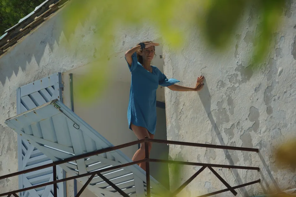 Femme en robe de coton bleu debout à un balcon, main en visière et paume posée sur un mur crépi blanc, à Porquerolles.