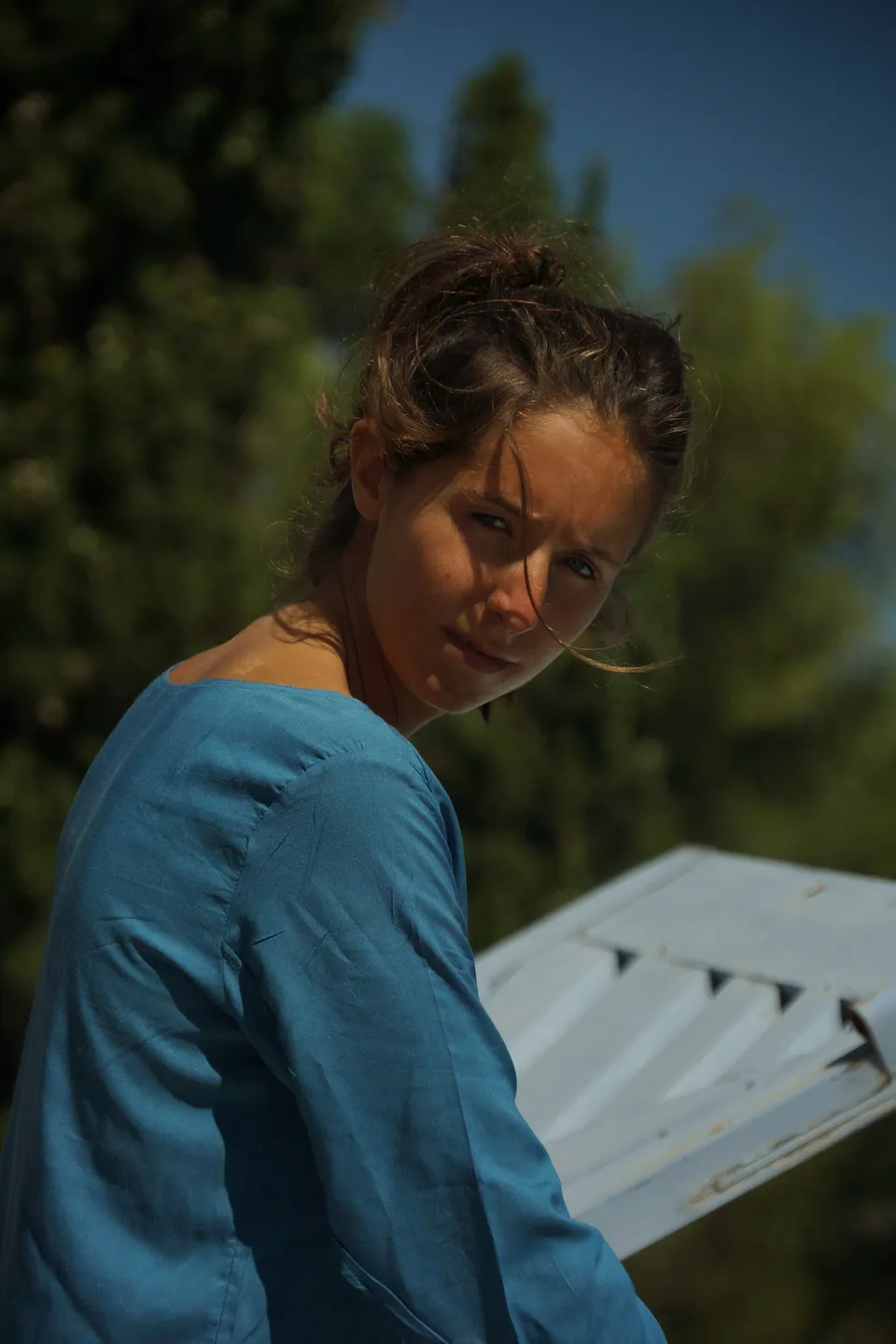 Portrait de trois quarts dos d'une femme en robe de coton bleu, épaule dégagée, regard vers l'objectif devant des pins, à Porquerolles.