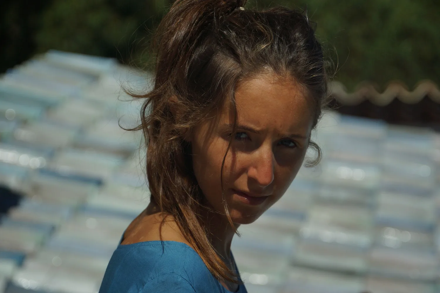 Portrait serré d'une femme en robe de coton bleu, cheveux au vent devant un toit de tuiles claires, à Porquerolles.