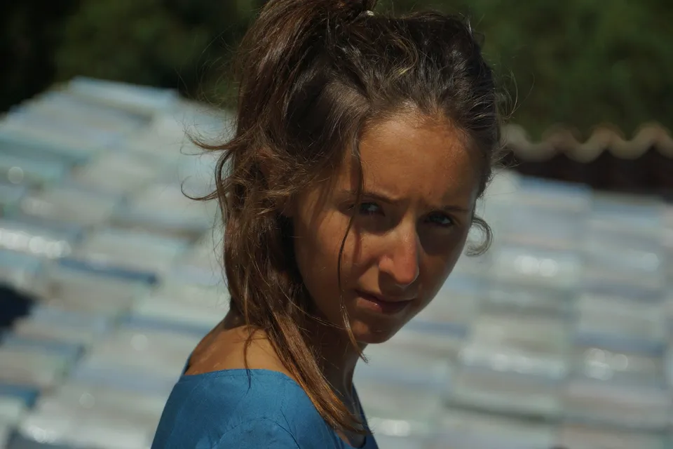 Portrait serré d'une femme en robe de coton bleu, cheveux au vent devant un toit de tuiles claires, à Porquerolles.