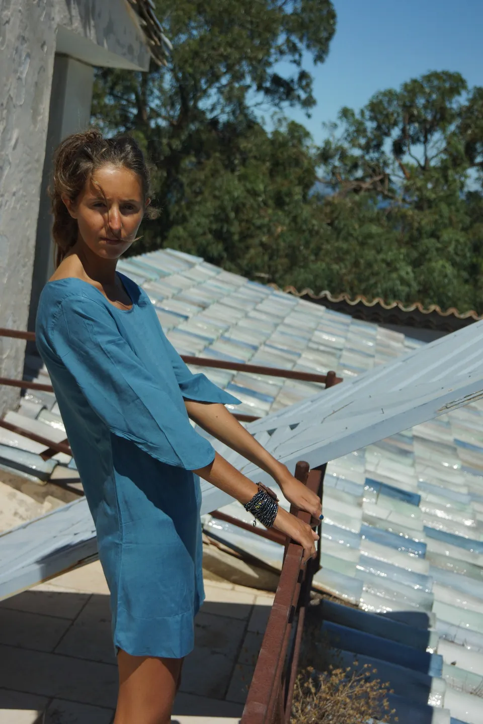 Femme en robe de coton bleu debout à une rambarde de fer, main sur un volet blanc, toit de tuiles derrière, à Porquerolles.