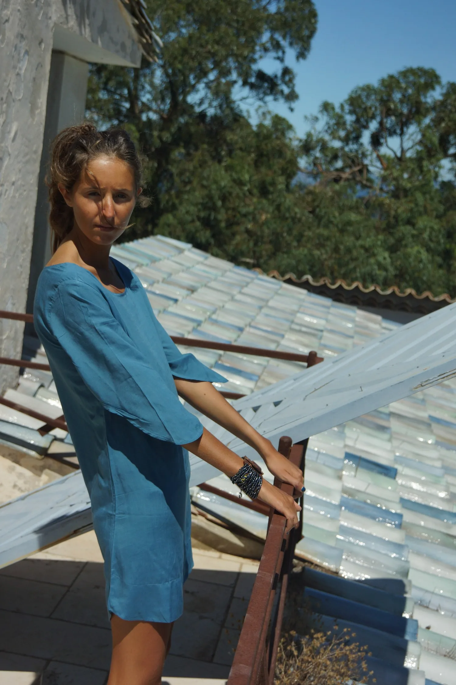 Femme en robe de coton bleu debout à une rambarde de fer, main sur un volet blanc, toit de tuiles derrière, à Porquerolles.