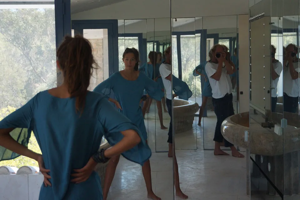 Femme de dos en robe de coton bleu, mains aux hanches, devant un mur de miroirs qui répète sa silhouette, à Porquerolles.
