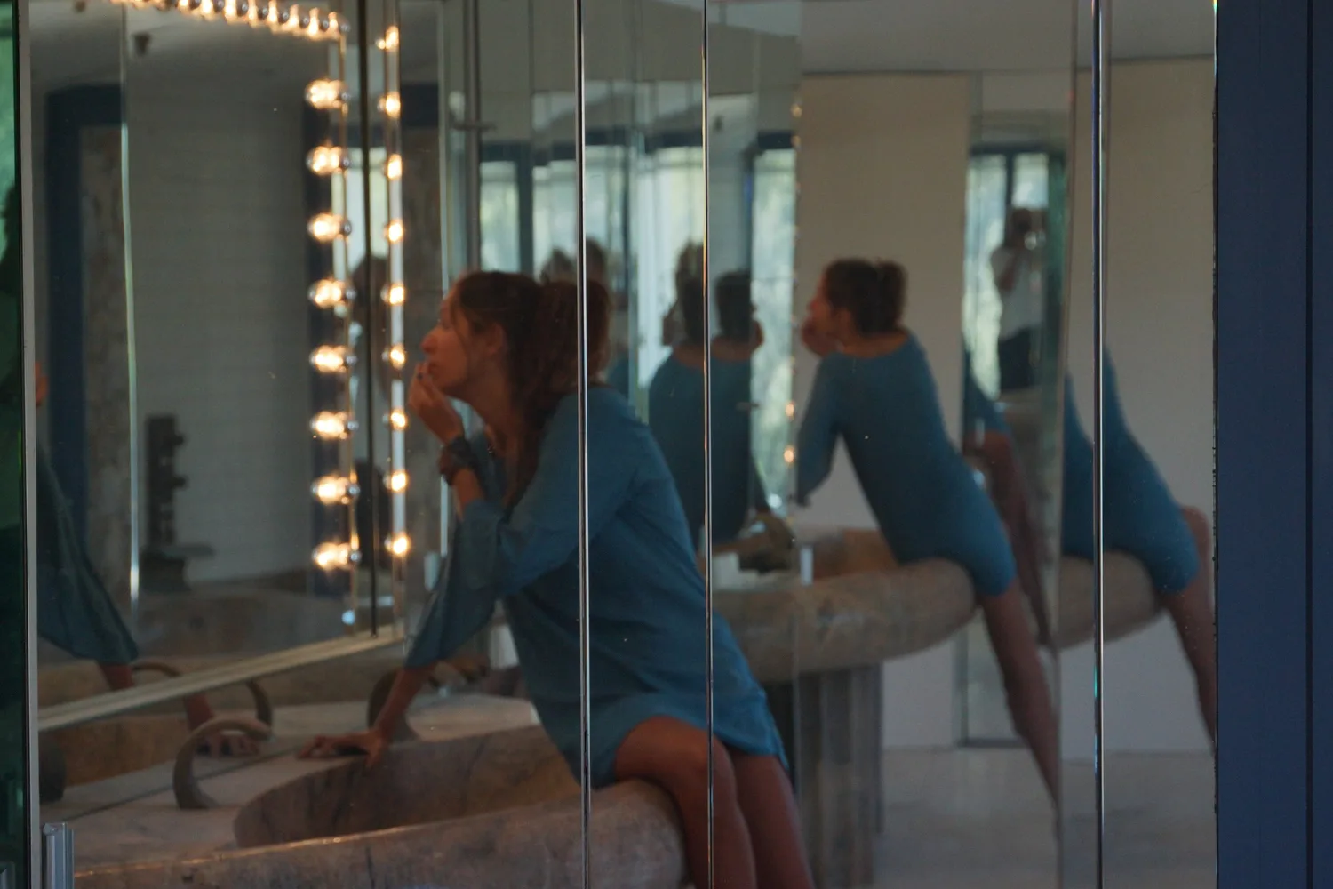 Femme en robe de coton bleu assise sur une vasque de pierre devant un miroir bordé d'ampoules, reflets en enfilade, à Porquerolles.
