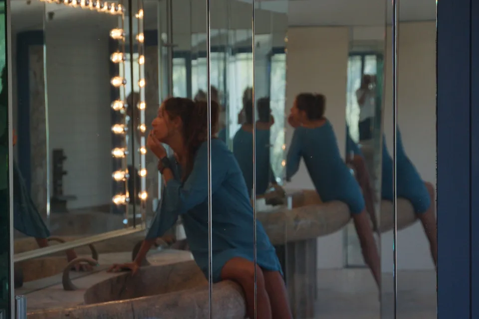 Femme en robe de coton bleu assise sur une vasque de pierre devant un miroir bordé d'ampoules, reflets en enfilade, à Porquerolles.
