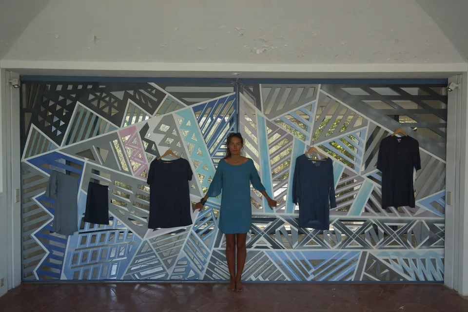 Femme en robe de coton bleu, bras ouverts devant une fresque géométrique bleue où sont suspendues cinq tuniques, à Porquerolles.