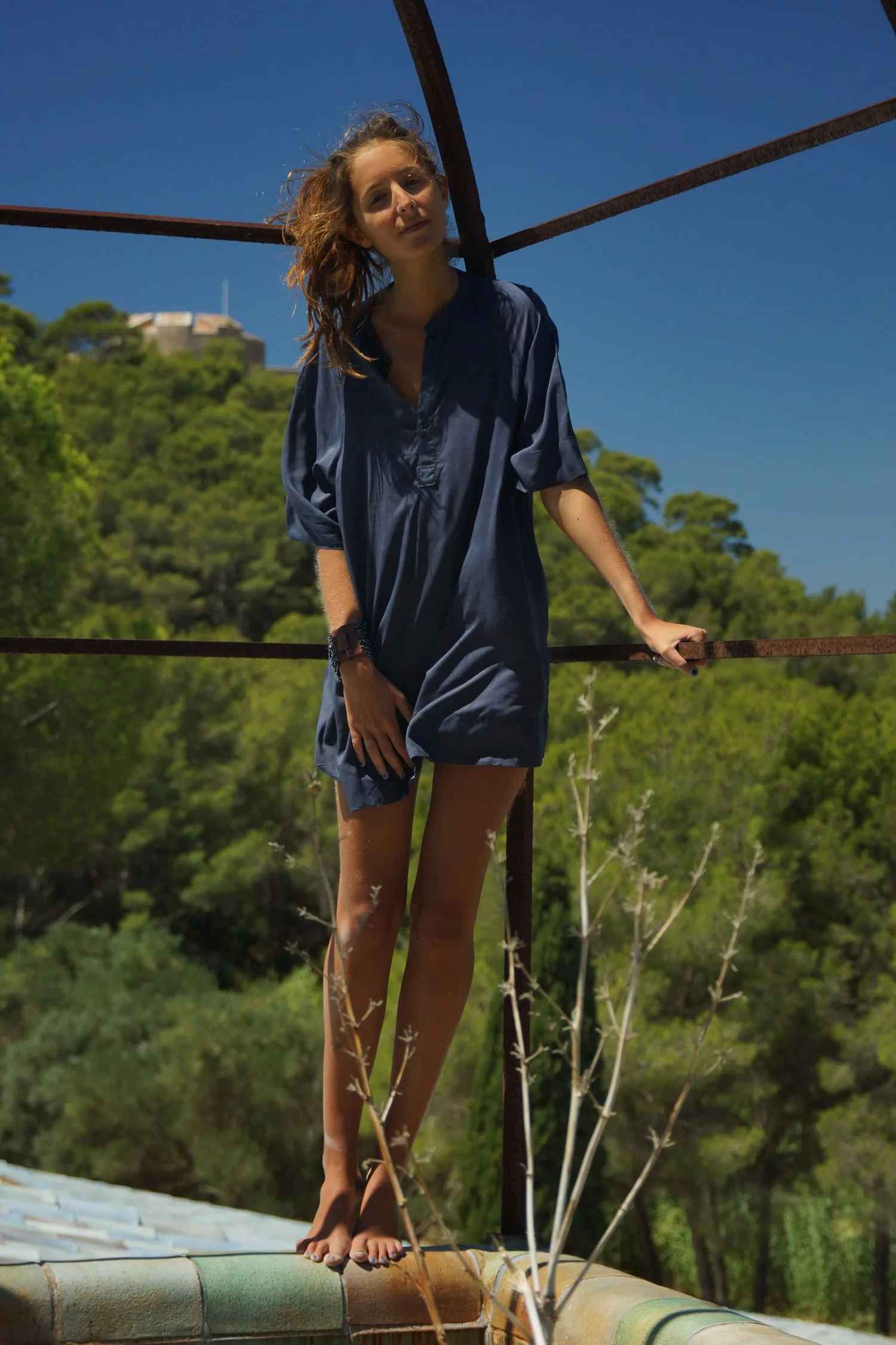 Femme en robe-chemise de coton bleu marine debout sur une banquette de céramique, pinède et fort au loin, à Porquerolles.