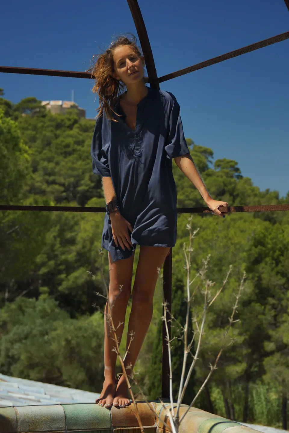 Femme en robe-chemise de coton bleu marine debout sur une banquette de céramique, pinède et fort au loin, à Porquerolles.