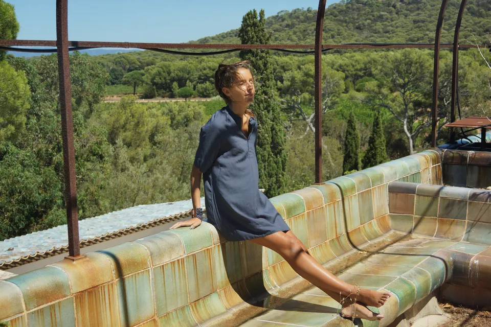 Femme en robe de coton bleu marine assise jambes tendues sur une longue banquette émaillée, cyprès et vallon derrière, à Porquerolles.