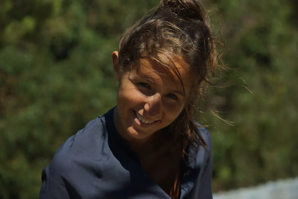 Portrait rapproché d'une femme souriante en robe de coton bleu marine, mèches au vent devant un feuillage sombre, à Porquerolles.