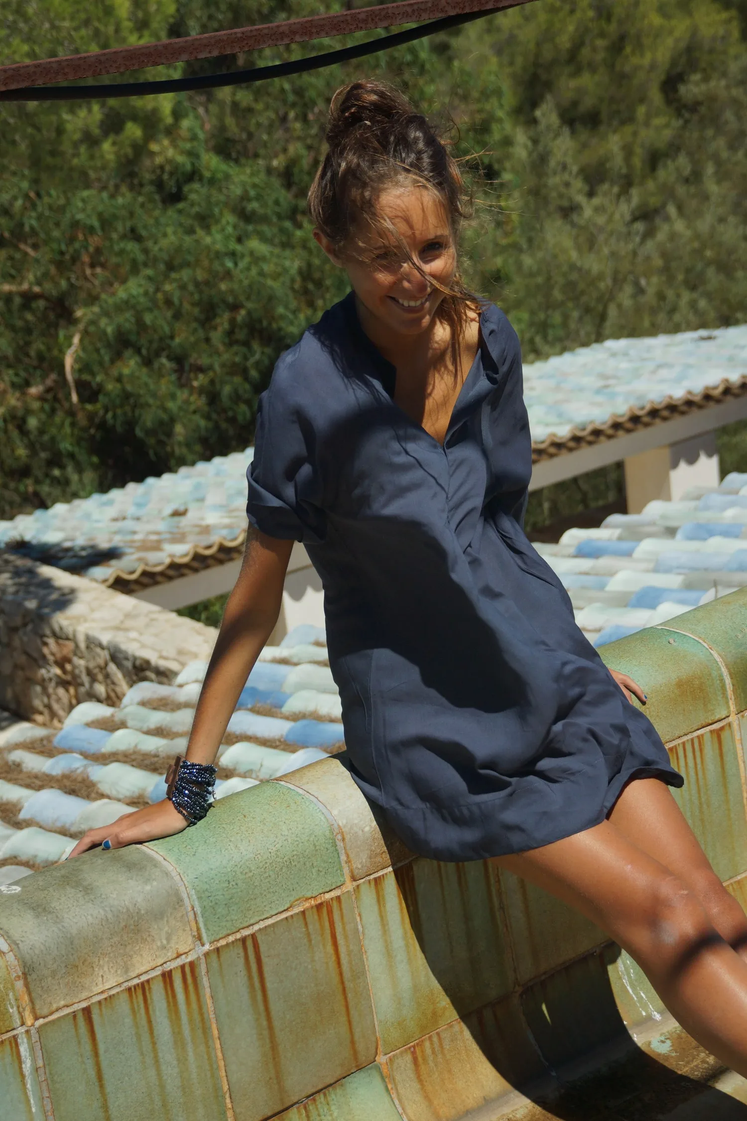 Femme en robe-chemise de coton bleu marine assise sur une banquette émaillée devant des toits de tuiles claires, à Porquerolles.