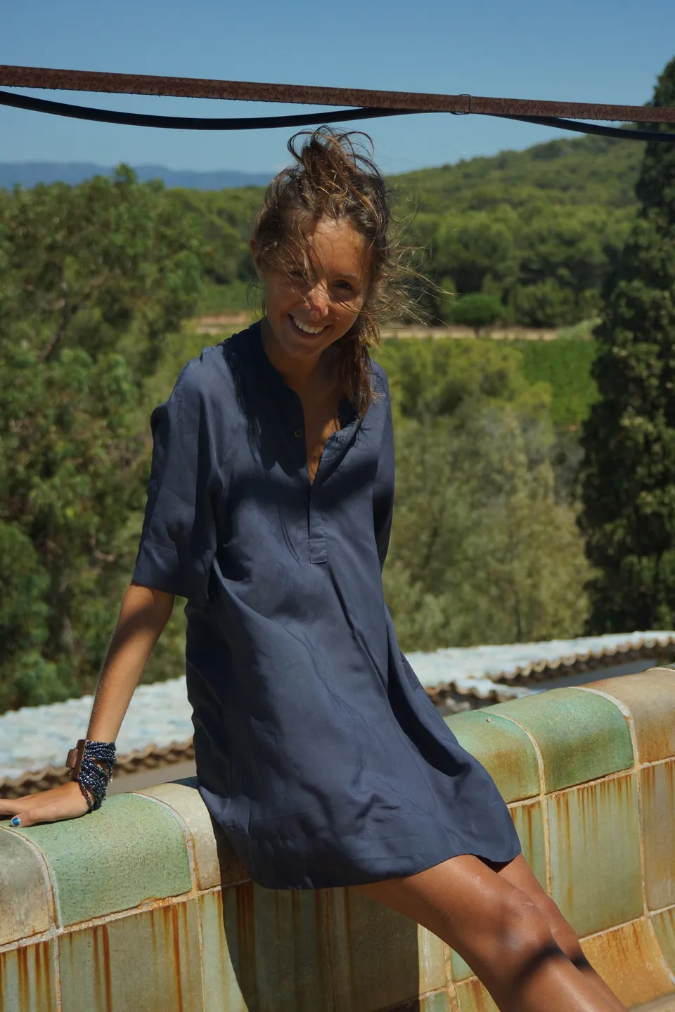 Femme en robe de coton bleu marine adossée à une banquette émaillée, vignes et collines derrière, à Porquerolles.