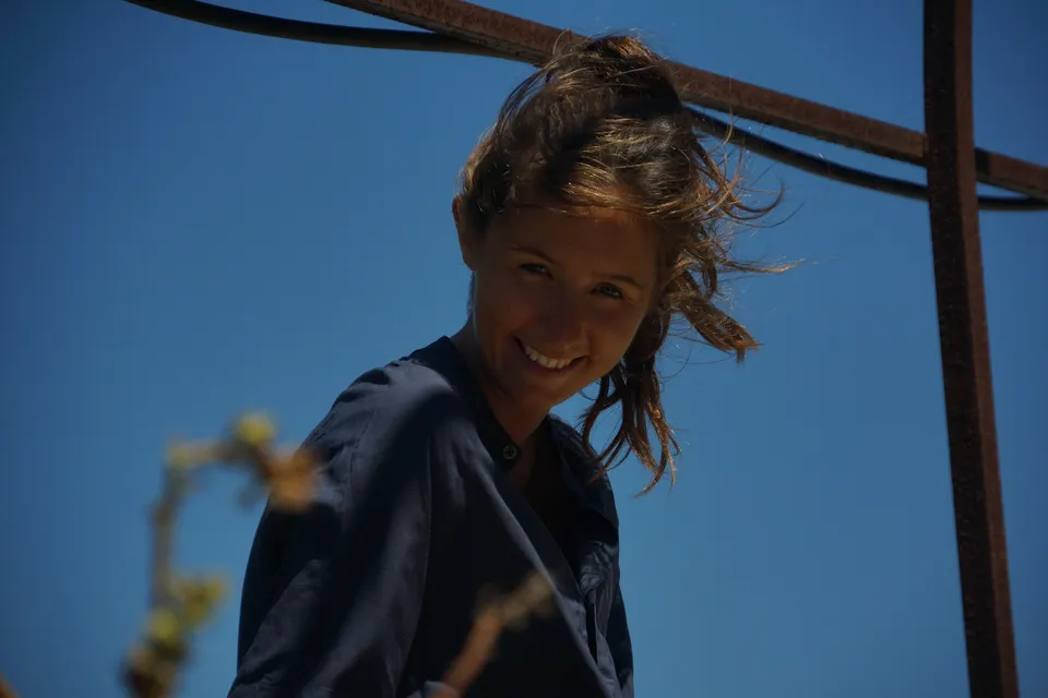 Portrait en contre-plongée d'une femme souriante en robe de coton bleu marine sur ciel bleu, barres de fer rouillé au-dessus, à Porquerolles.