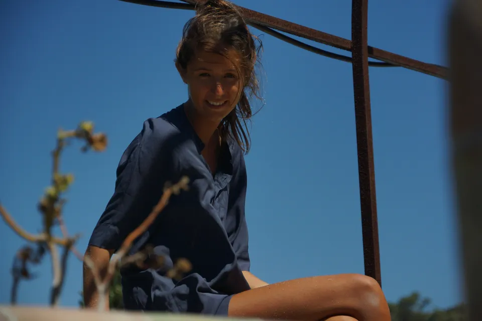 Femme en robe de coton bleu marine assise sous une pergola de fer, ciel bleu plein cadre et brindilles sèches en amorce, à Porquerolles.