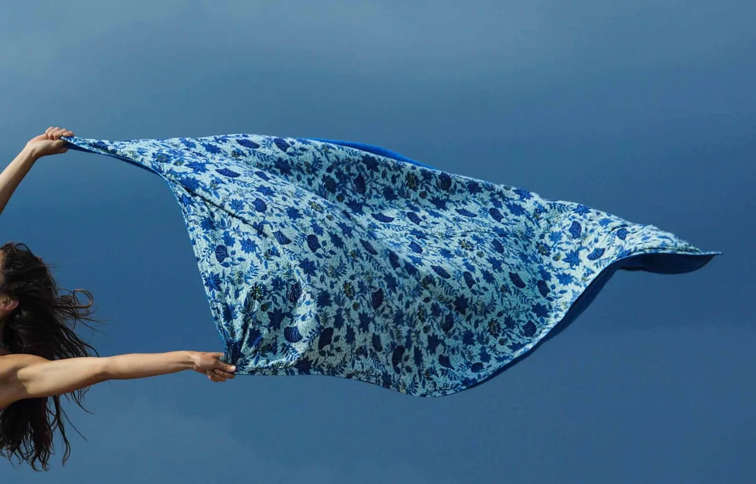 Femme bras levés déployant un grand foulard imprimé bleu dans le vent devant une mer sombre, à Porquerolles.